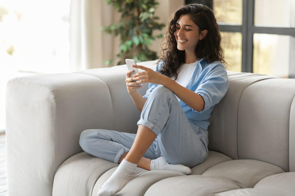Portrait of cheery young woman using smartphone, sitting on comfy sofa at home, copy space